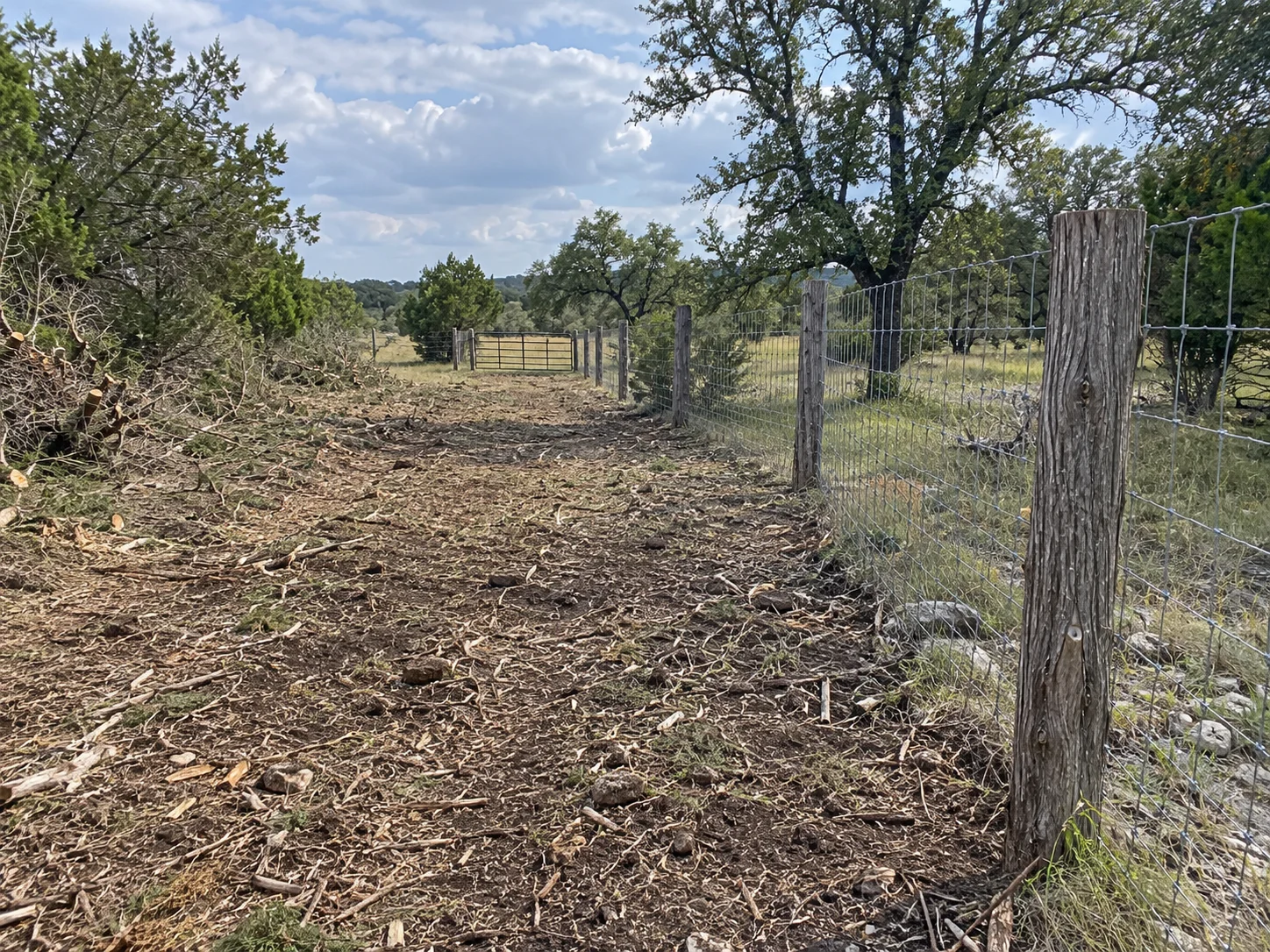 Cleared ranch access lane beside a wire fence with a metal gate ahead.