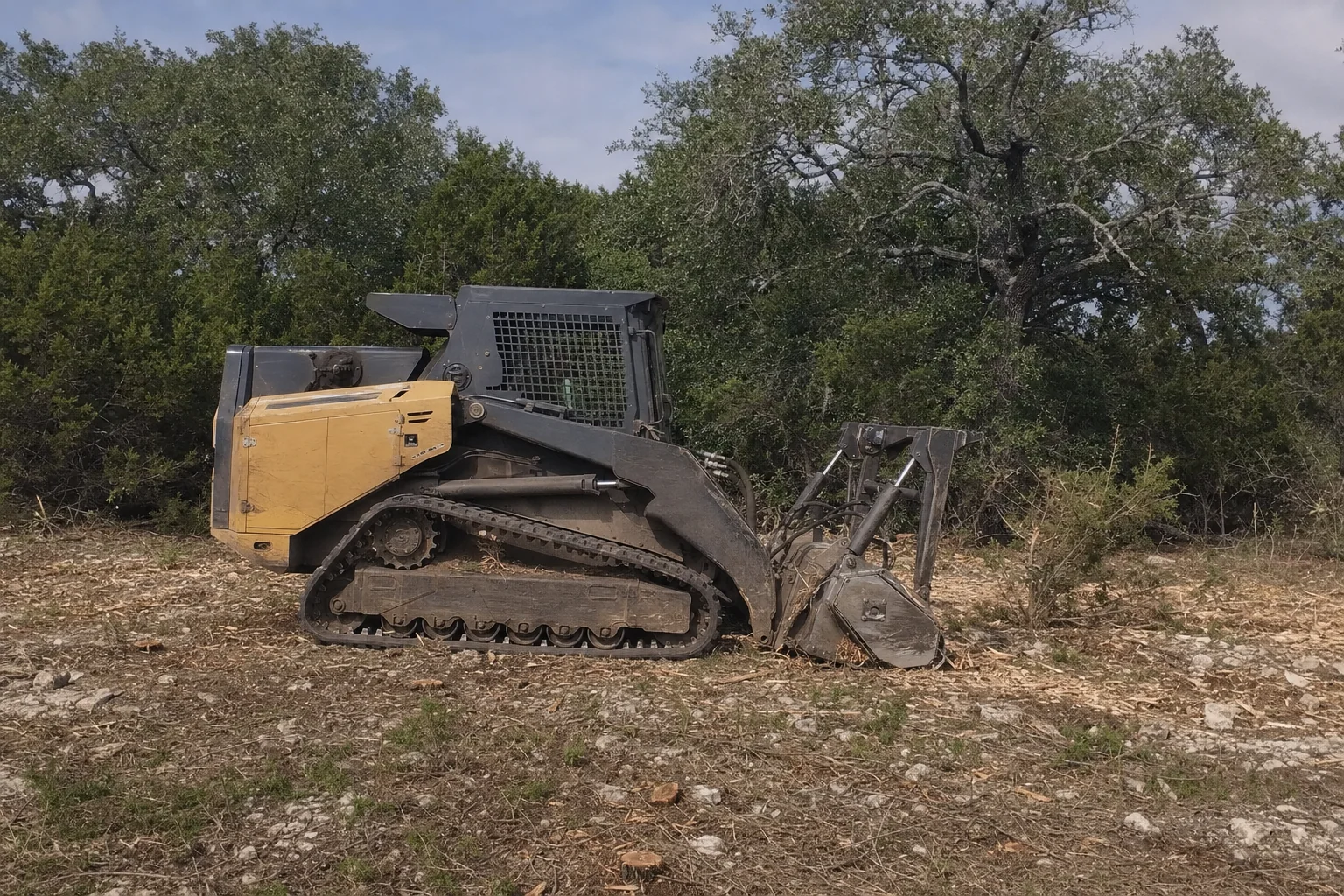 The mulcher rests on rocky ground beside cedar brush and retained oaks.
