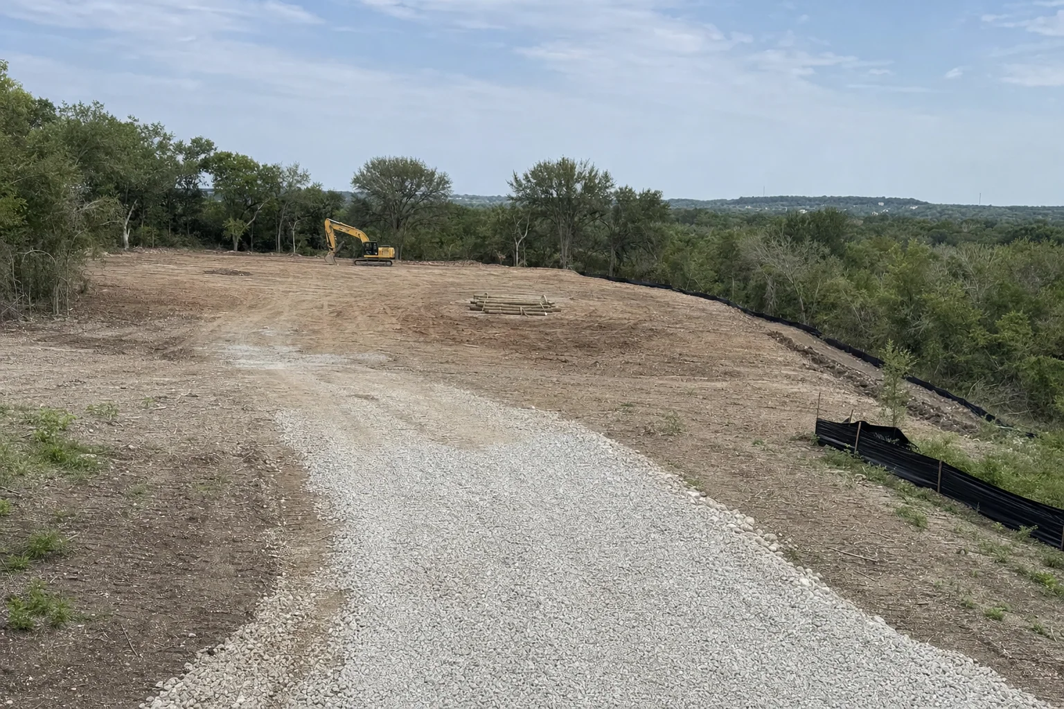 A gravel entrance leads past stacked logs toward a parked excavator.