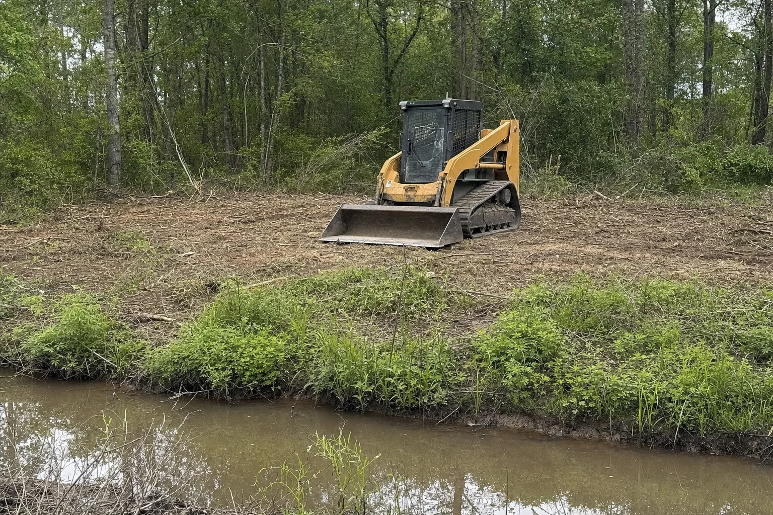 Across the muddy ditch, a loader sits before the remaining leafy woodland.