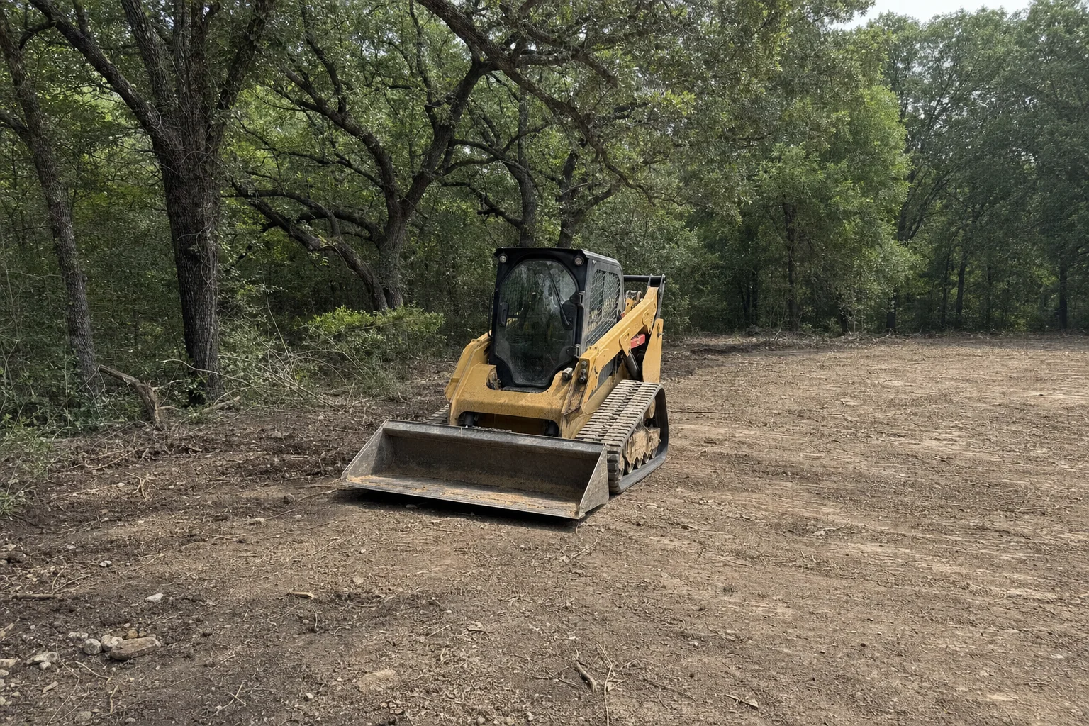 Oak limbs shade the loader at the edge of the cleared footprint.