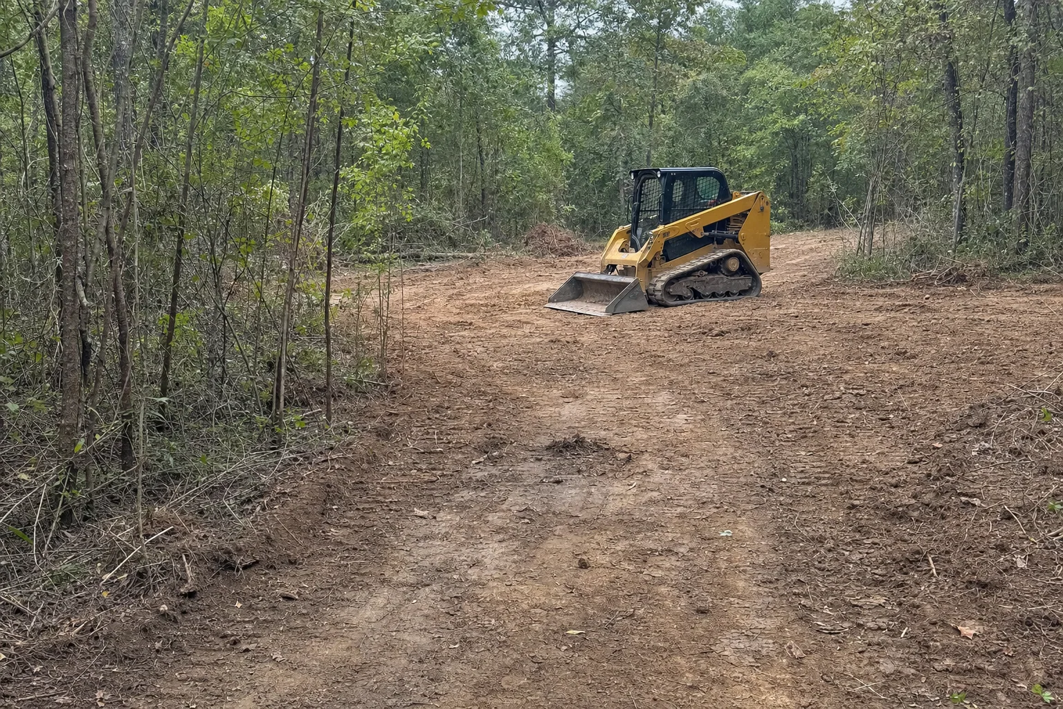 The woodland lane widens into open turning room around a parked loader.