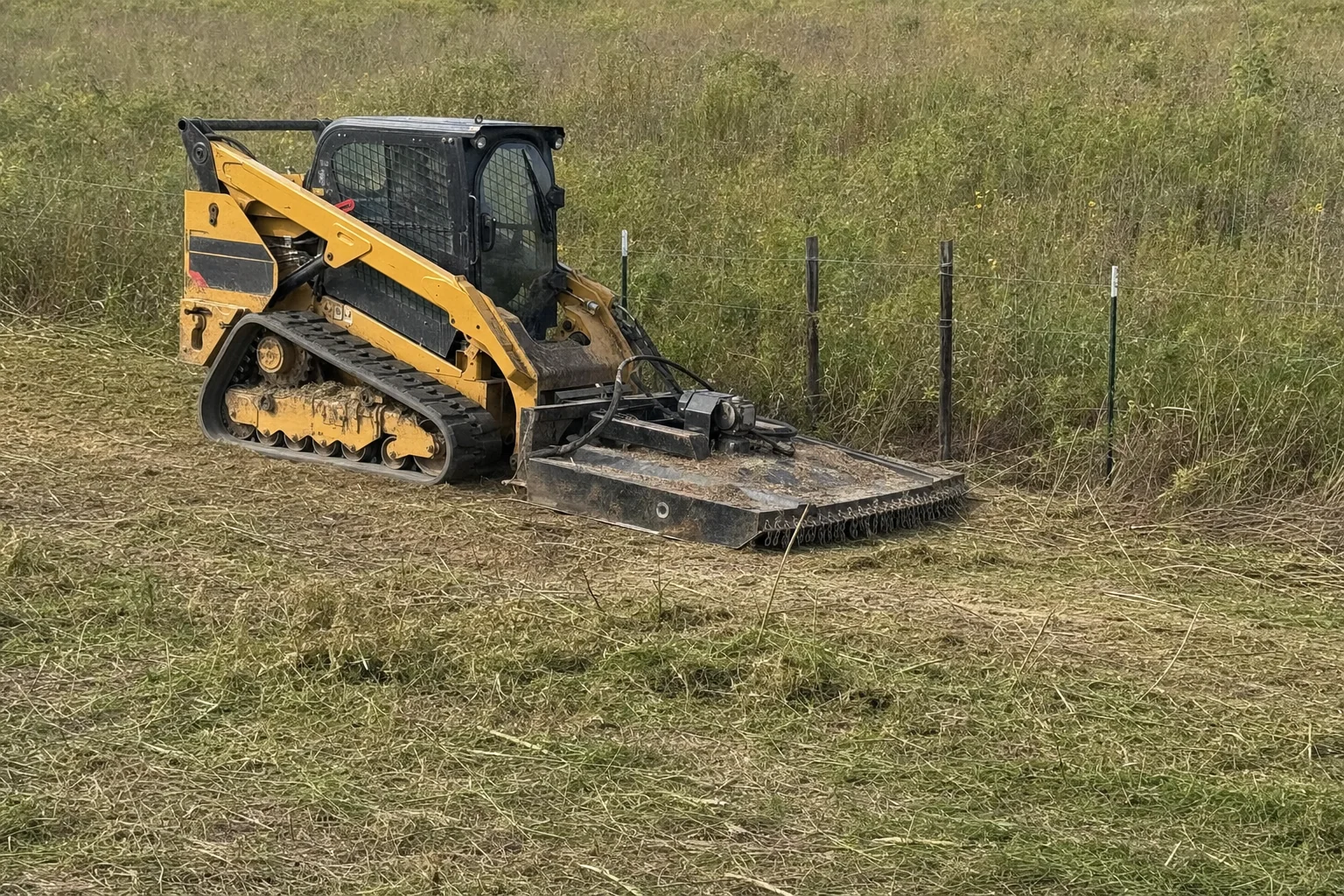 Coarse mowing cuttings lie below the wire fence and lowered rotary deck.