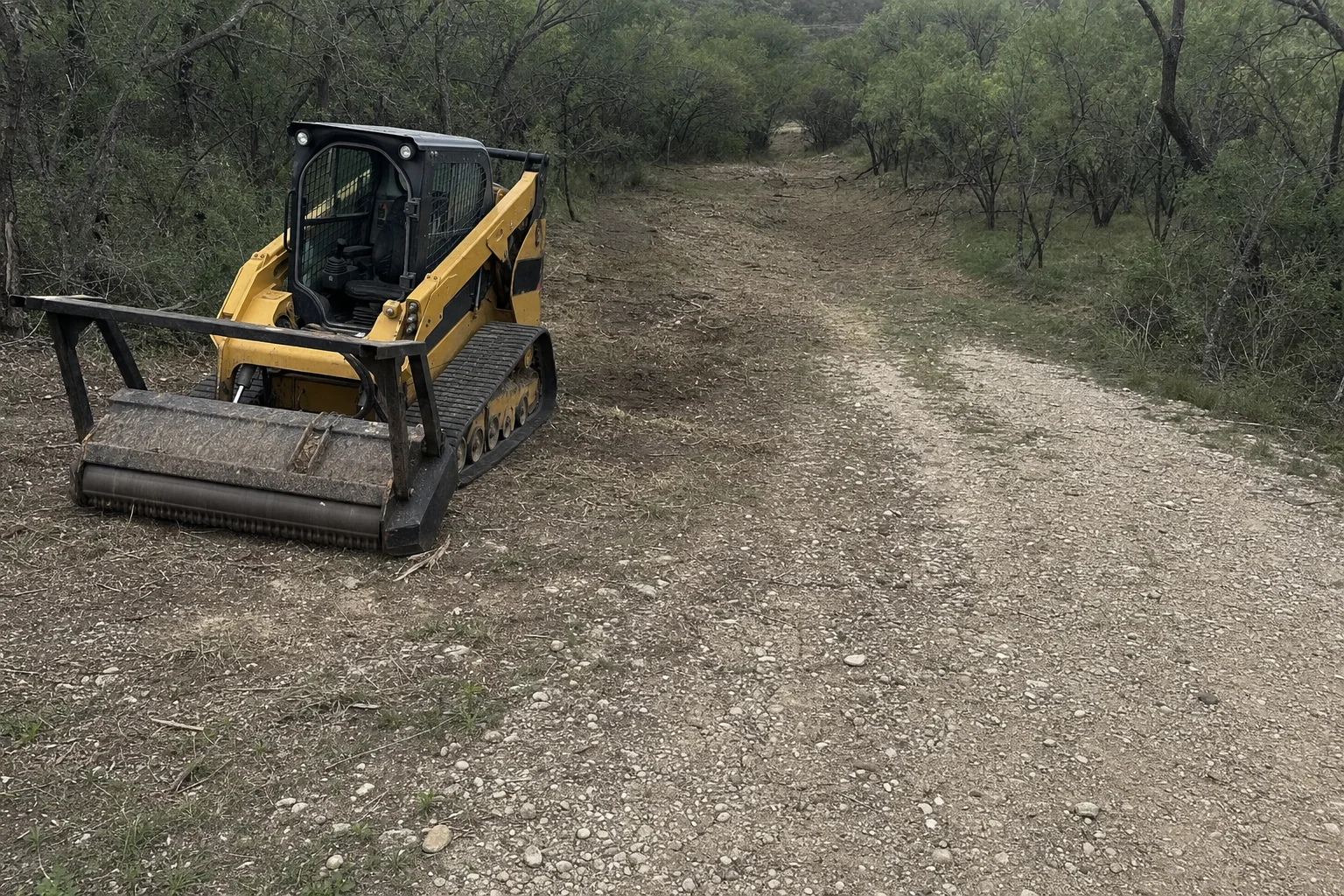 Gravel meets the opened brush corridor beside the parked loader.