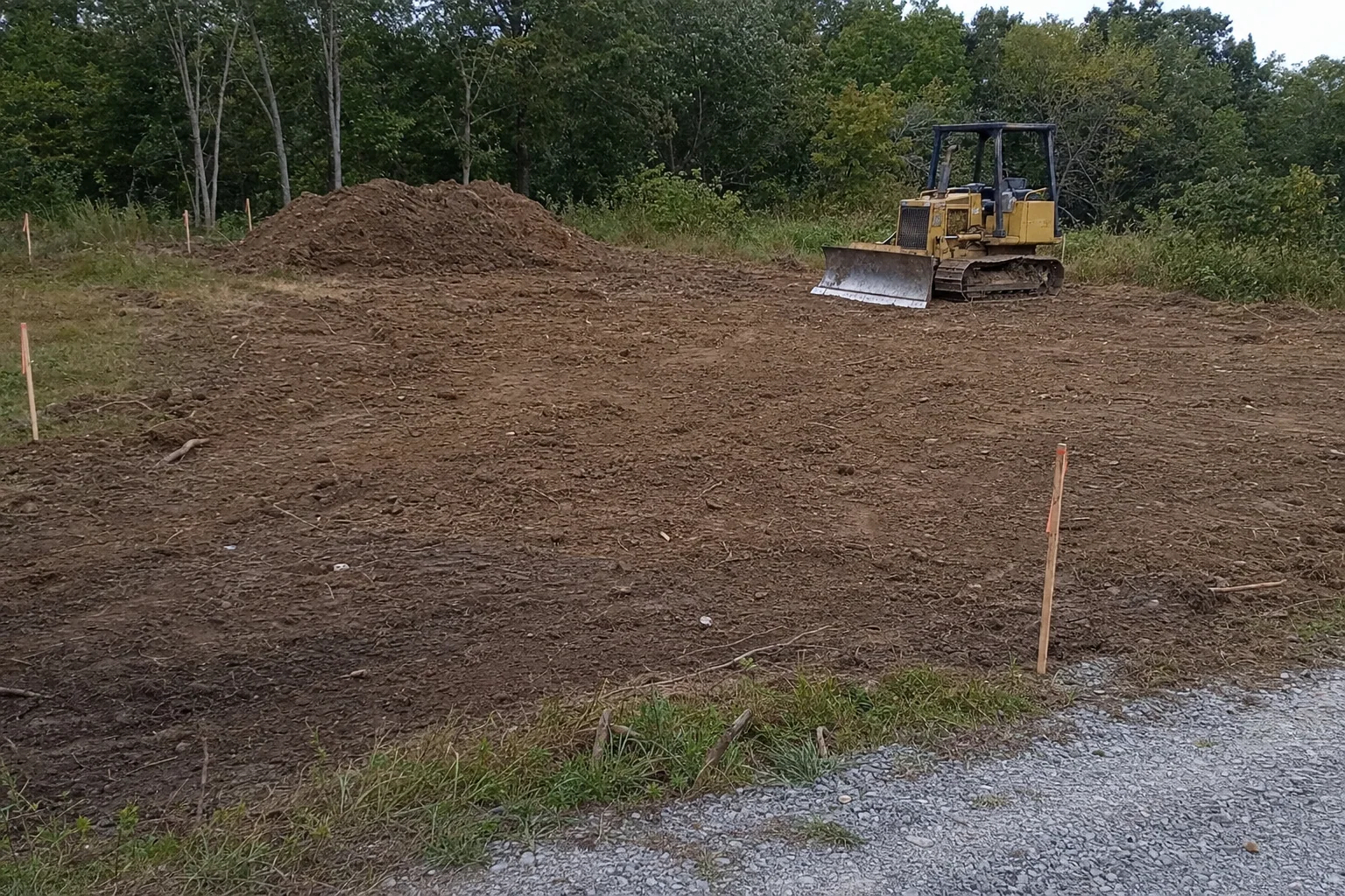 Survey stakes edge the rough footprint, with a dozer and soil pile behind.